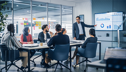 Several people sit at a conference table, looking at a person who is pointing to data on a large screen